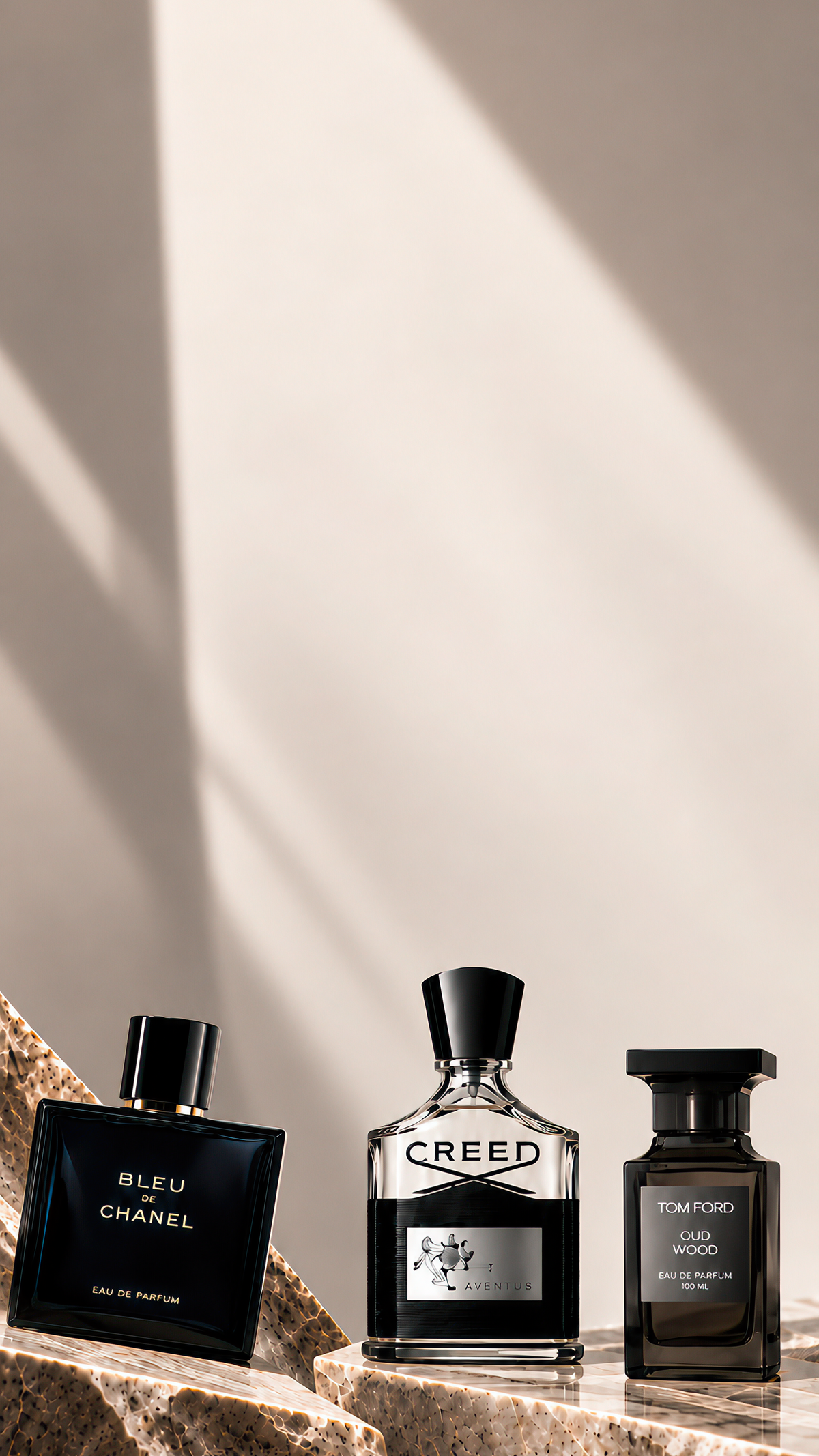 Three luxury fragrance bottles arranged on a stone slab against a warm beige wall with diagonal shadows. The bottles include Bleu Chanel, Creed, and Tom Ford, all in dark glass with black caps and silver-toned labels.